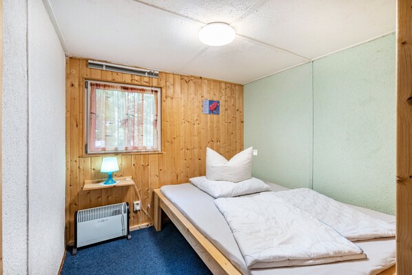 A small, single-bed room with a wooden headboard, a window with curtains, a bedside table with a lamp, and a radiator. - Bungalow '6 - Finnhütte' with Private Terrace and Shared Garden (Niesky)