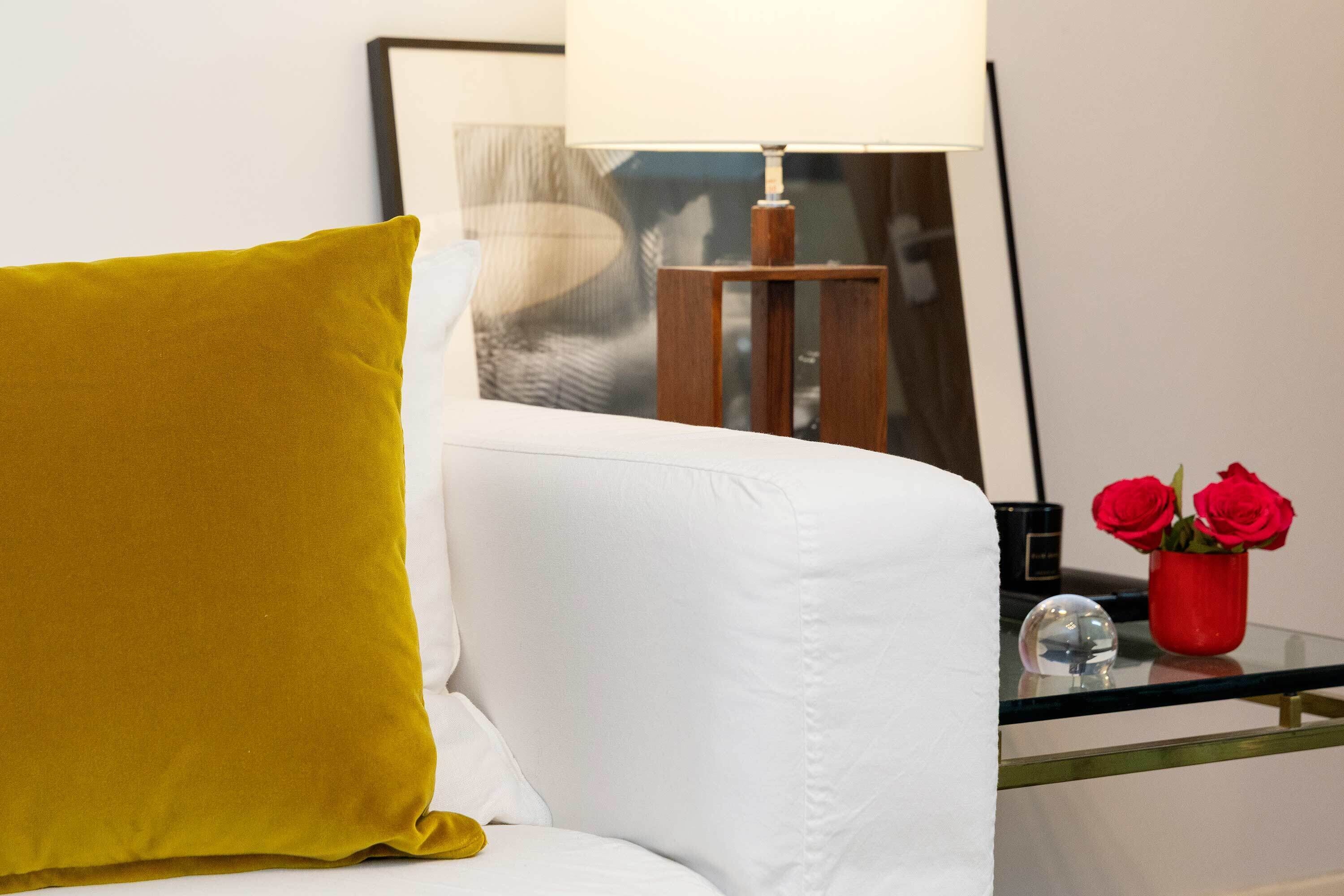 A white sofa with a mustard-colored pillow, a glass table with a red vase of roses, and a framed picture on the wall.