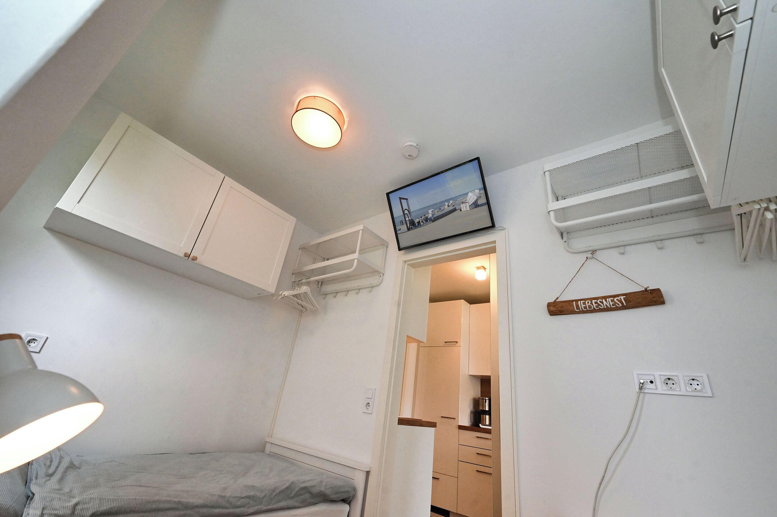 Un dormitorio compacto con cama, escritorio, silla, lámpara colgante, ventilador de techo, cuadro enmarco y una pequeña zona de cocina.