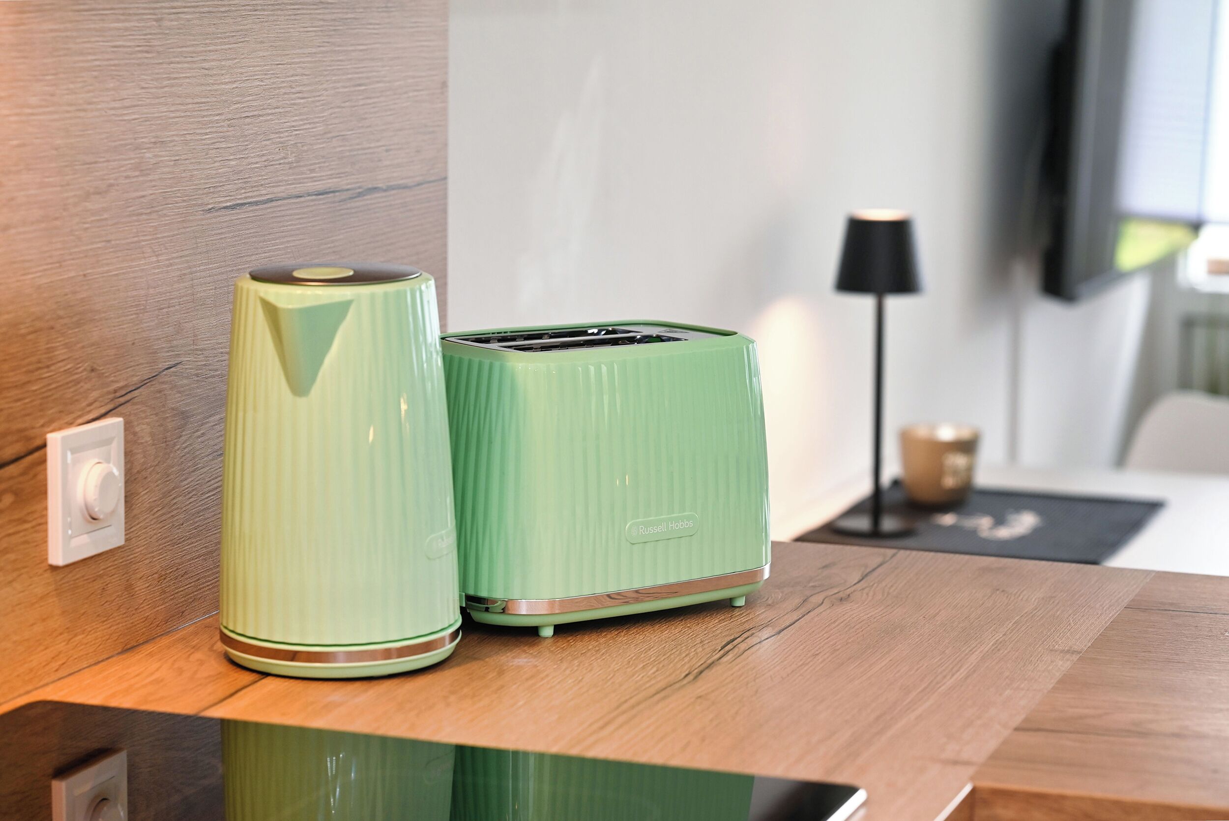 Two green appliances on a wooden countertop, a lamp in the background, and a wall switch.