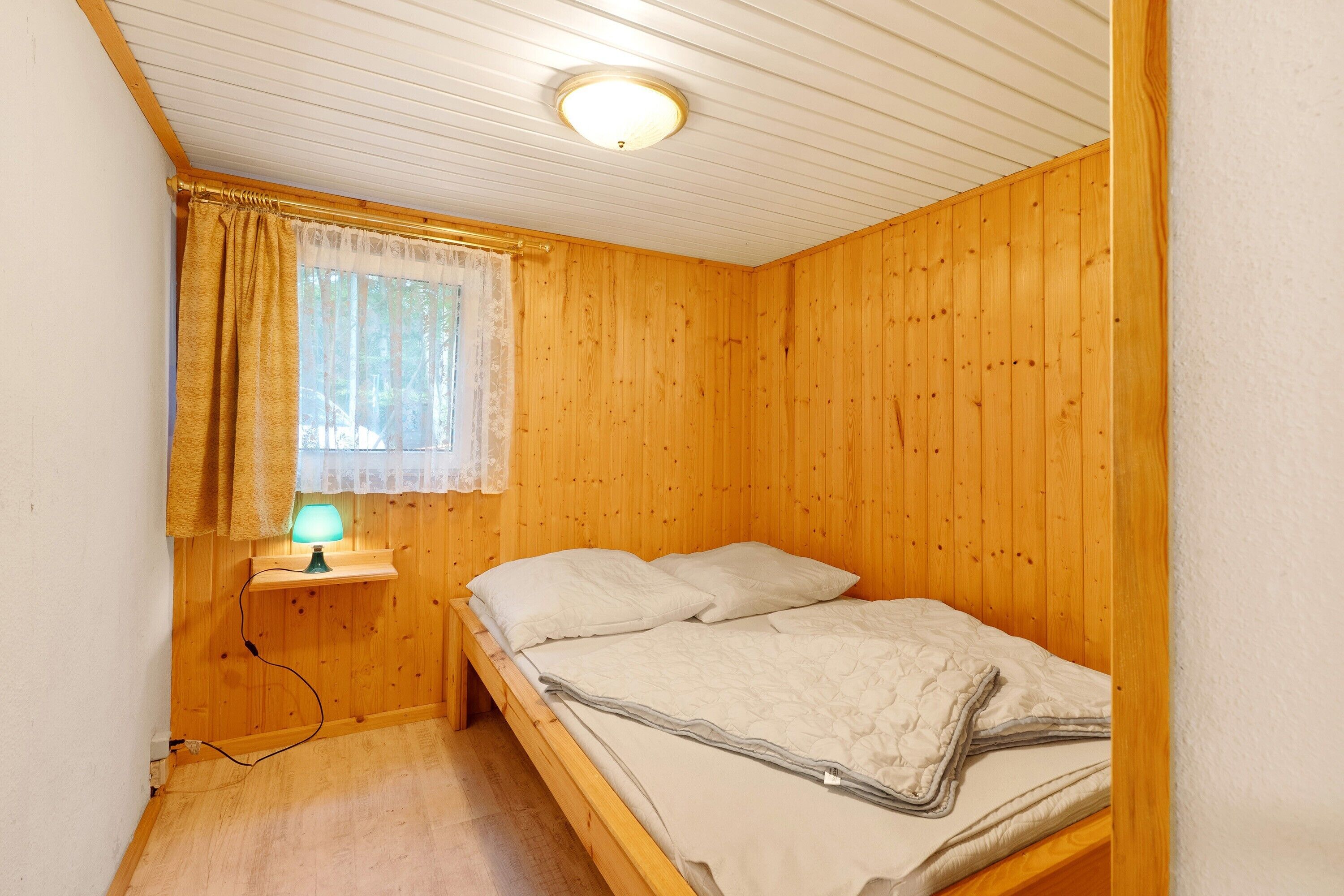 Ein Einzelbett mit Holz-Kopfstück, ein Nachttisch mit Lampe und ein Fenster mit Vorhängen.