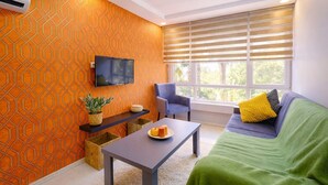 A modern living room with a geometric patterned wall, a flat-screen TV, a sofa with cushions, a chair, a small table with a plant and a pumpkin, and a window with blinds.