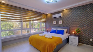 A modern bedroom with a large bed, a bedside table with a lamp, and a view of greenery outside.
