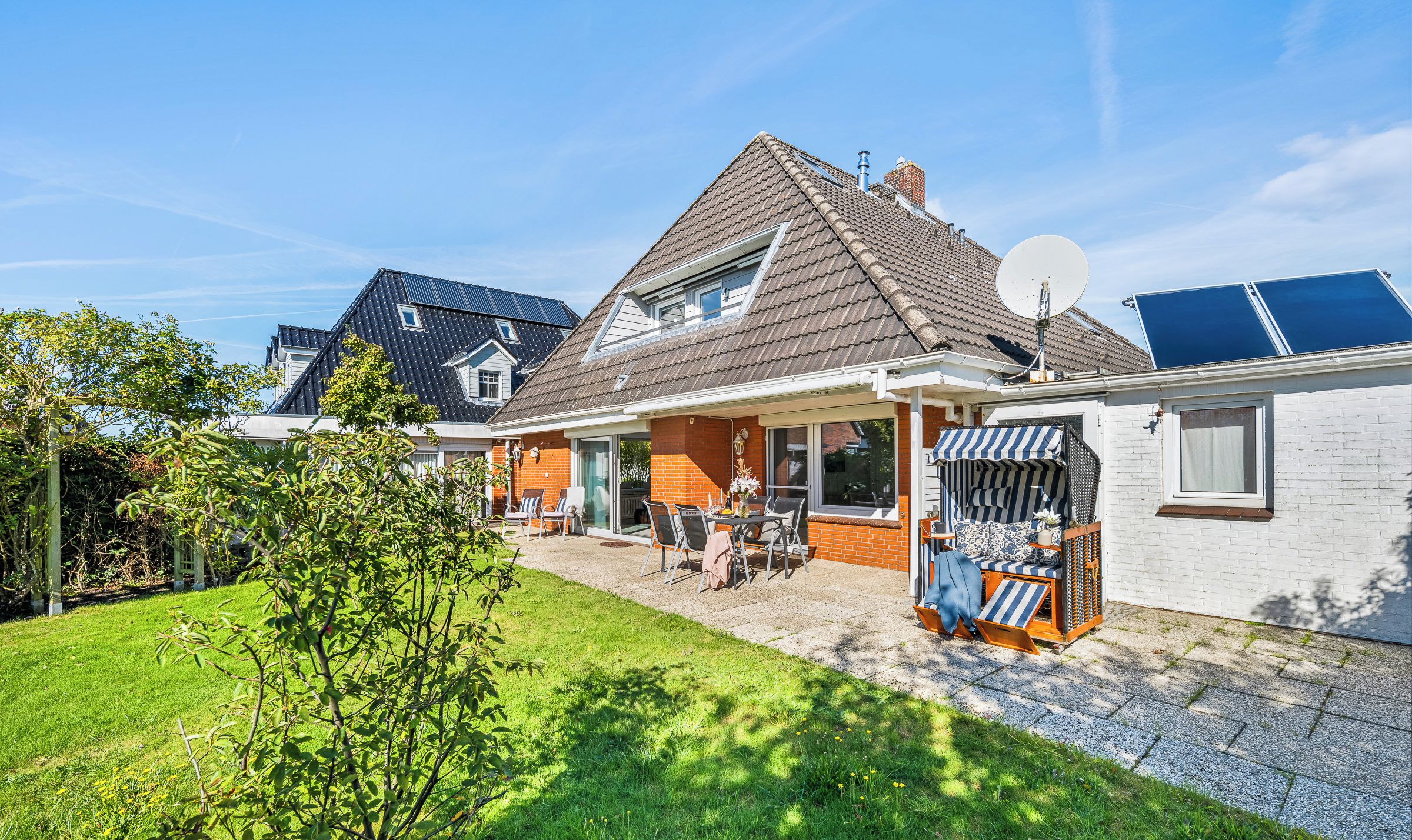 Une maison d’habitation avec un toit en pente, des panneaux solaires, une antenne parabolique et une terrasse aménagée avec du mobilier d’extérieur.