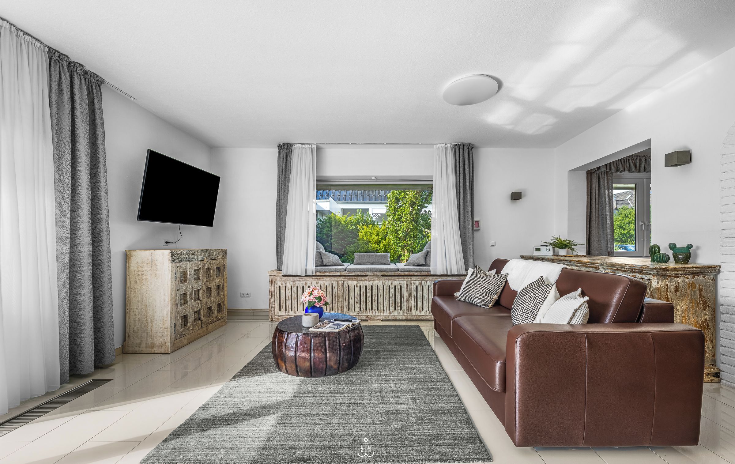Un salón moderno con un sofá de cuero marrón, una mesa de centro redonda de madera, un televisor de pantalla plana montado en la pared y un amplio ventanal con cortinas.