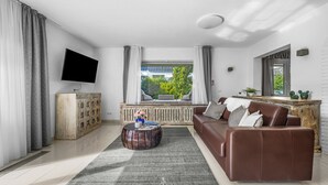 Un salón moderno con un sofá de cuero marrón, una mesa de centro redonda de madera, un televisor de pantalla plana montado en la pared y un amplio ventanal con cortinas.