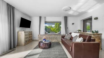 Un salón moderno con un sofá de cuero marrón, una mesa de centro redonda de madera, un televisor de pantalla plana montado en la pared y un amplio ventanal con cortinas.