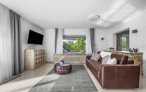 Un salon moderne avec un canapé en cuir marron, une table basse ronde en bois, un téléviseur à écran plat fixé au mur et une grande fenêtre avec des rideaux.