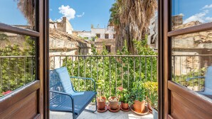 A balcony with a chair and potted plants, overlooking a historic building with a tiled roof and palm trees.