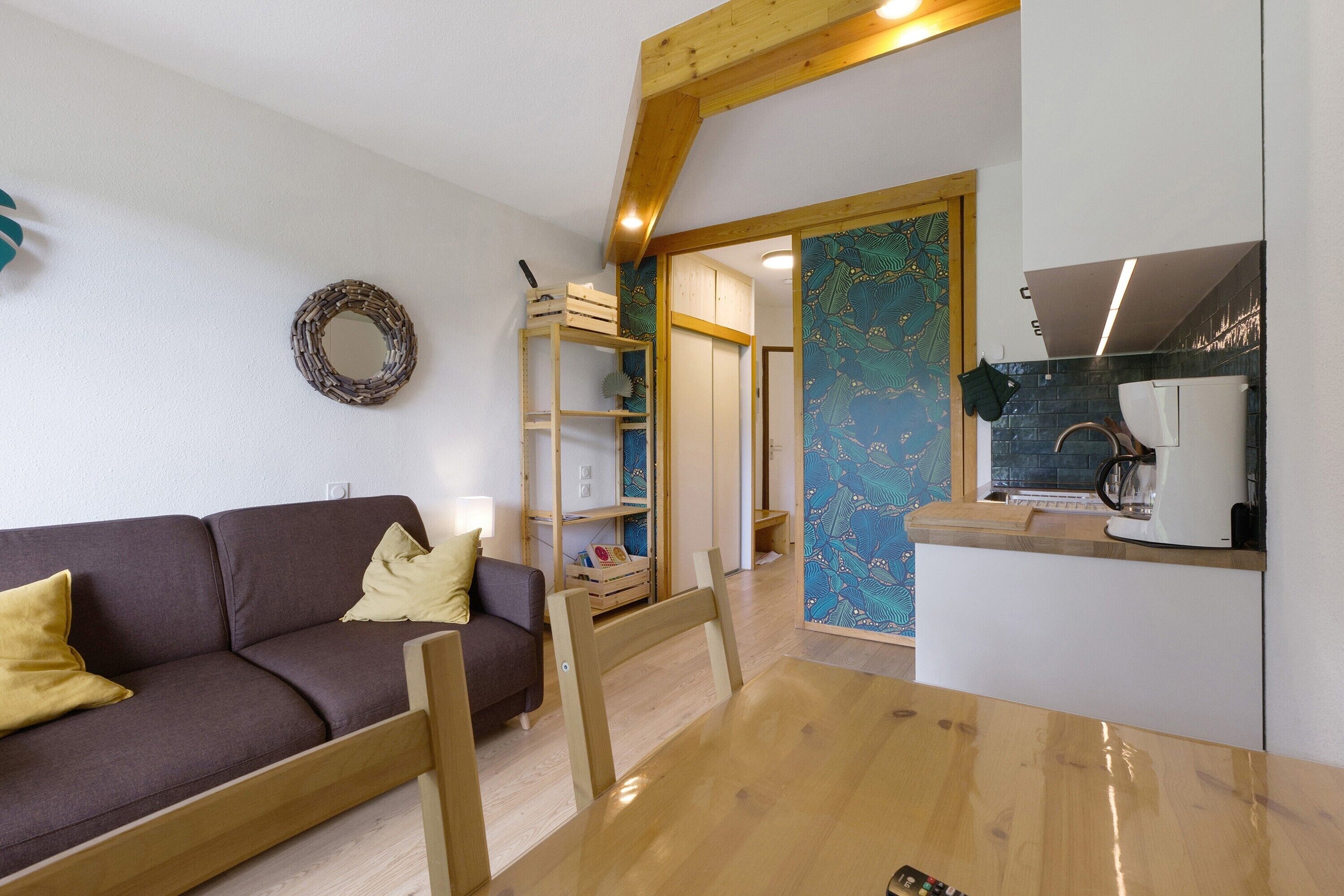 A modern interior with a wooden dining table, a sofa, a kitchen area, and a patterned wall.
