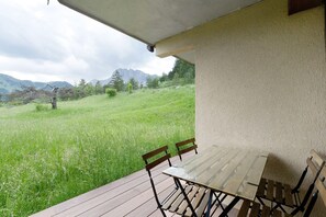 En udendørs terrasse med et bord og stole, med udsigt til en græsplæne og bjerge.