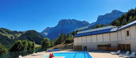 A swimming pool with lounge chairs and a building with solar panels, set against a mountainous backdrop.