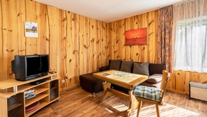 Une pièce aux murs lambrissés, avec un téléviseur posé sur un meuble, un canapé, une table avec un set de table et un radiateur.