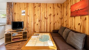 Une pièce aux murs lambrés, avec un téléviseur posé sur un meuble en bois, un canapé et une table basse sur laquelle est posée une carte.