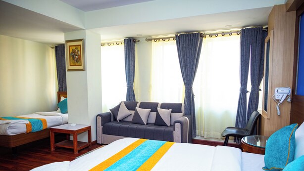 A hotel room with a large bed, a sofa, a small table, and a chair. There is a window with curtains and a painting on the wall. - Hotel Sara Inn (Kathmandu)