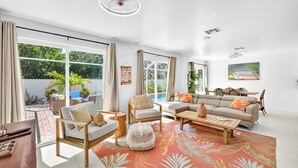 A spacious living room with a large window, a sofa set, a coffee table, and a dining area with chairs. - Lower Villa Nature View-Heated Pool/Block to Sand (Fort Myers Beach)