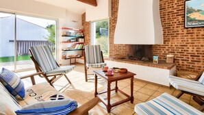A cozy living room with a brick fireplace, striped chairs, a bookshelf, and a large window overlooking a garden. - 700m from the beach, garden, 4 adults 2 children (La Guérinière)