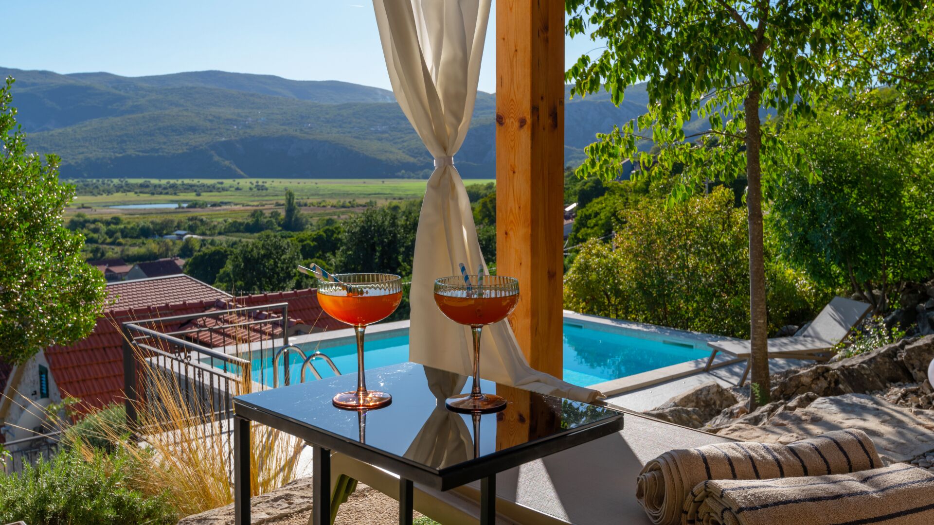 Un tavolo vicino alla piscina con due bevande, vista su una piscina e un paesaggio montuoso.