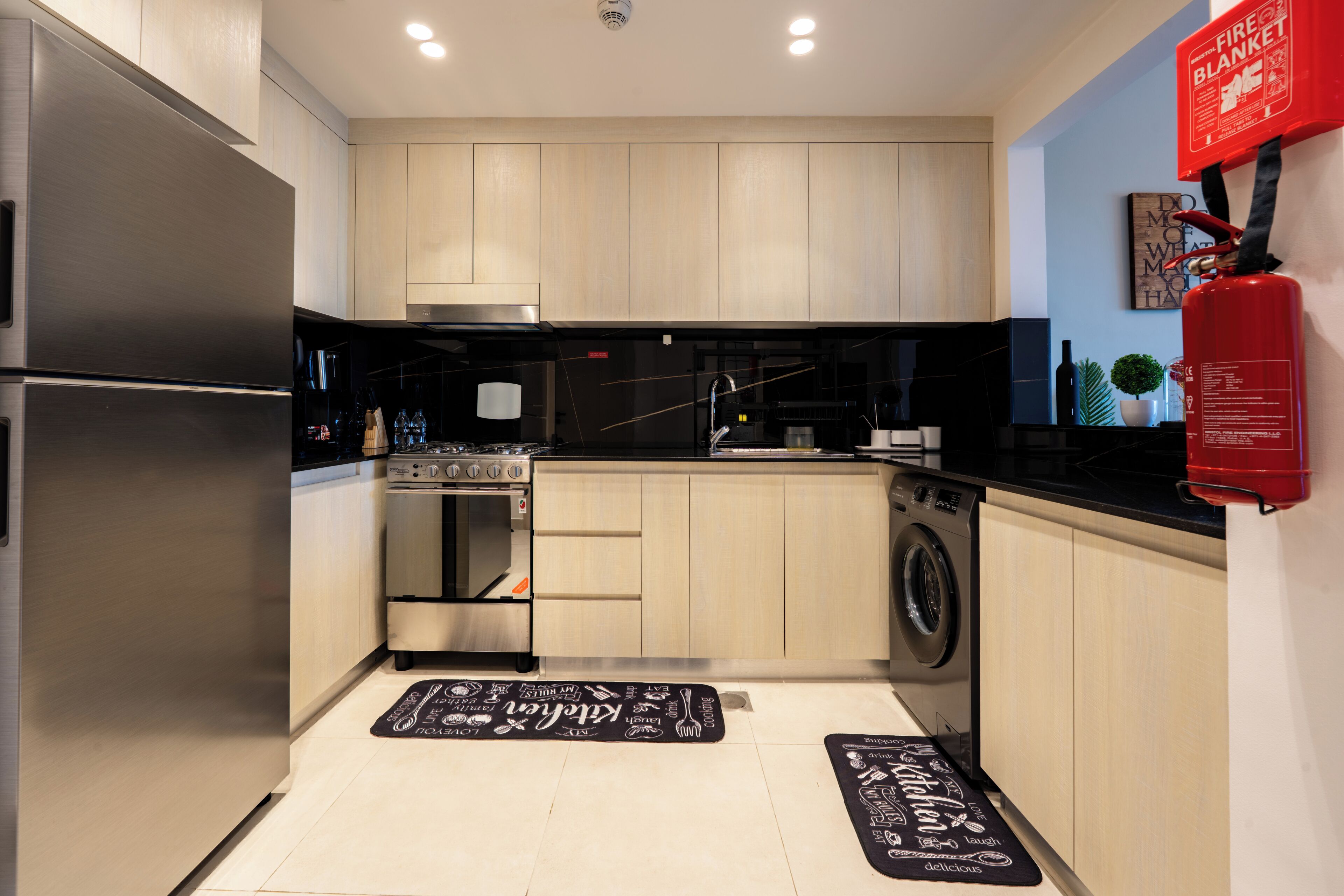 A modern kitchen with stainless steel appliances, wooden cabinets, a washing machine, and a fire extinguisher.