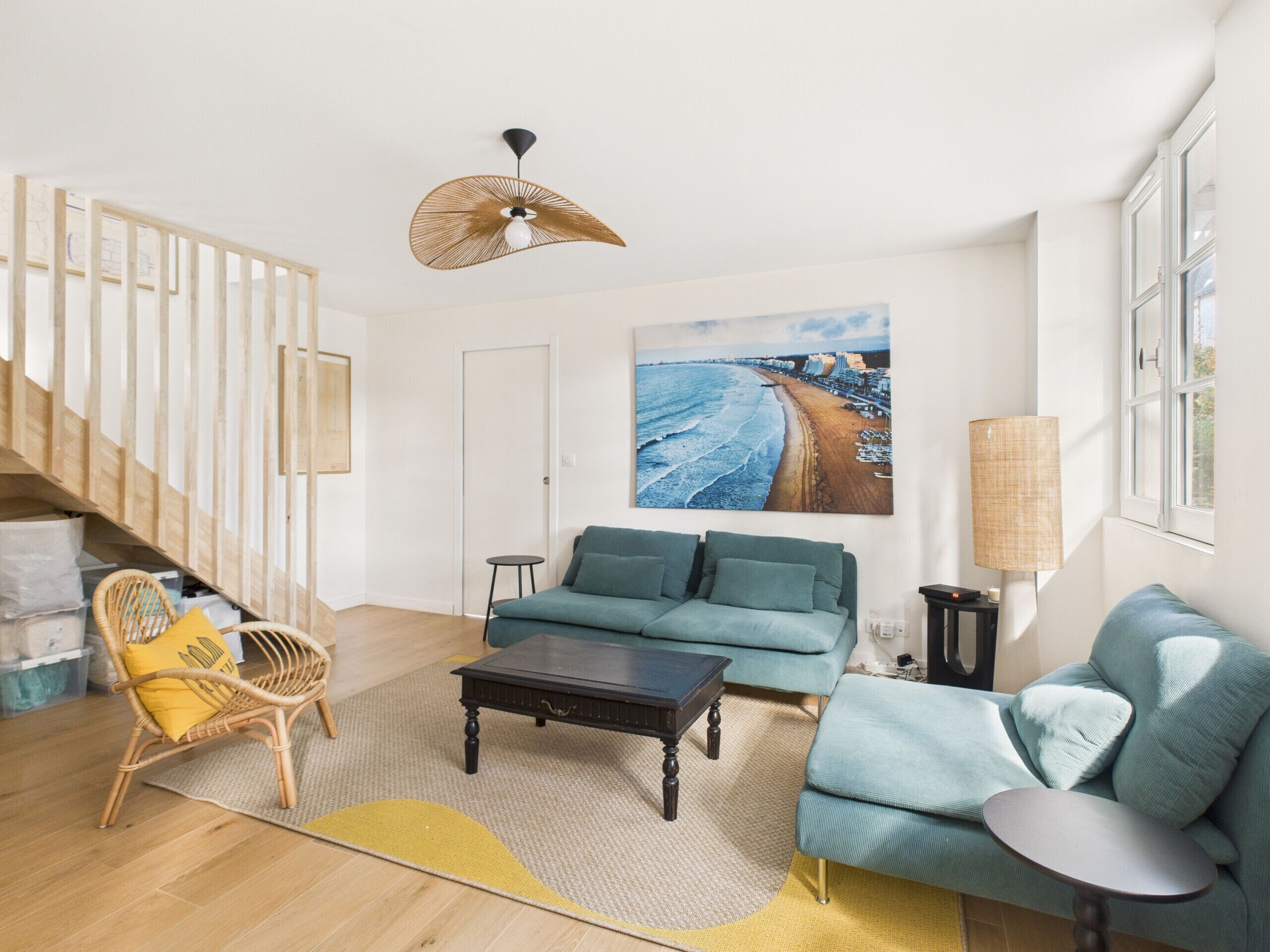 A modern living room with a sofa, coffee table, and a chair. There is a painting of a beach on the wall, a lamp, and a staircase leading to another room.