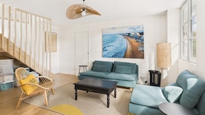 A modern living room with a sofa, coffee table, and a chair. There is a painting of a beach on the wall, a lamp, and a staircase leading to another room. - 250m from beach - house for 6 adults/2 kids (Pornichet)