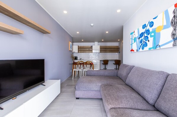 A modern living room with a grey sectional sofa, a flat-screen TV on a white stand, and a kitchen in the background with wooden cabinets and a white backsplash. - Pool View 1 Bed Apt T2-720 (Chalong)