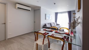 A modern dining area with a glass table, white chairs, and a grey sofa in the background. - Pool View 1 Bed Apt T2-720 (Chalong)