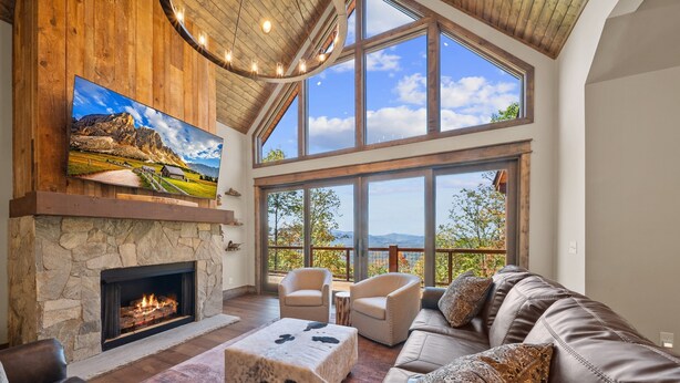 A spacious living room with a stone fireplace, leather furniture, and large windows offering a mountain view. - Golden Sunsets at Eagles Nest - hot tub, view, community amenities (Banner Elk)