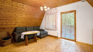 Ein Zimmer mit Holzboden, einer Couch, einem kleinen Tisch, einem Korbstuhl und einem großen Fenster mit Vorhängen.