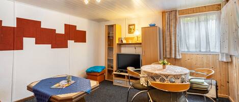 Quarto com uma mesa redonda, cadeiras, televisão, estante e janela com cortinas.