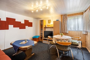 Une pièce comprenant une table ronde, des chaises, un téléviseur, une bibliothèque et une fenêtre avec des rideaux.