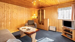 Una habitación con paneles de madera, un sofá, una mesa con un control remoto, un televisor sobre un mueble bajo y una ventana con cortinas.