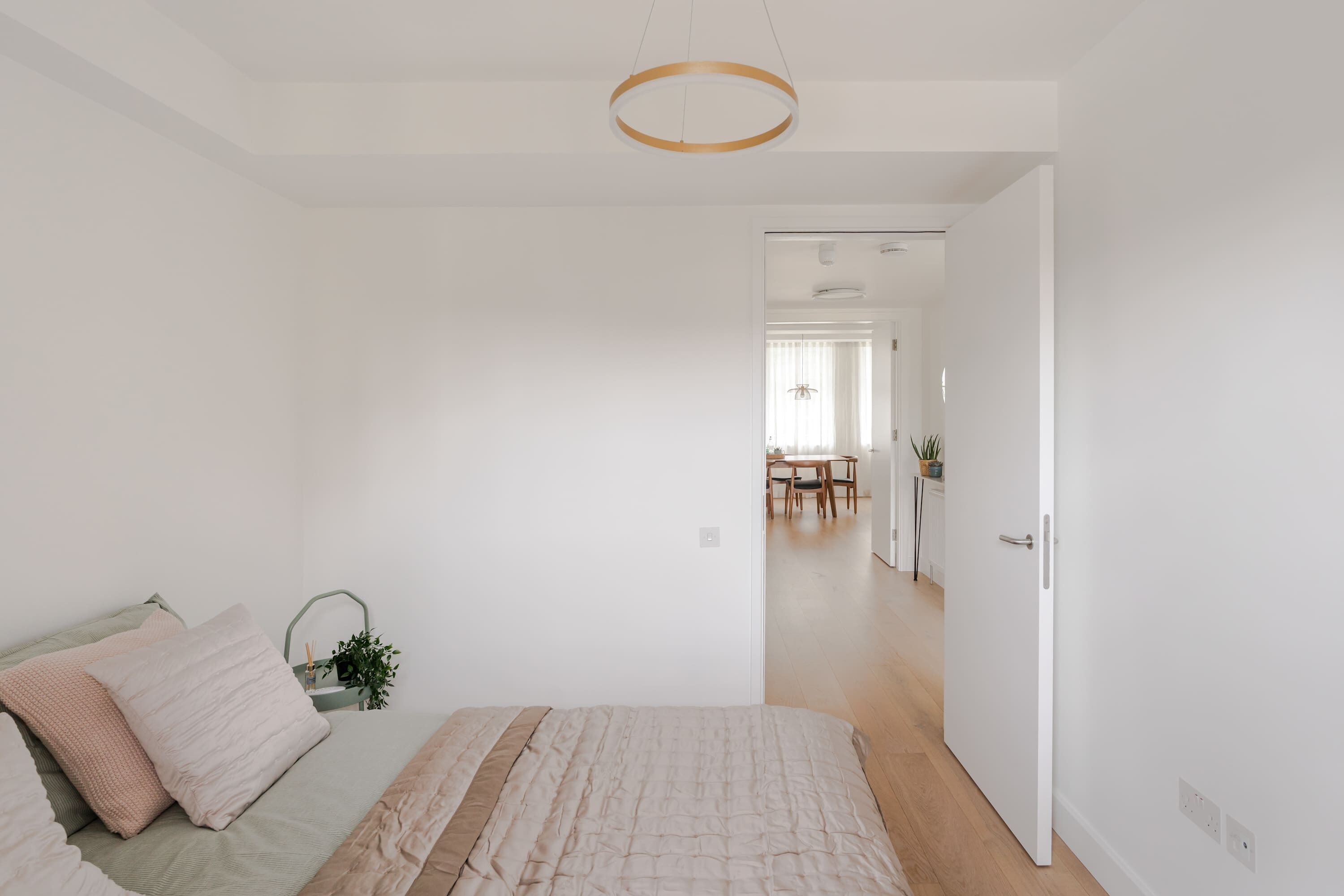 A bedroom with a bed, a bedside table, a hanging light fixture, and a view into the dining area.