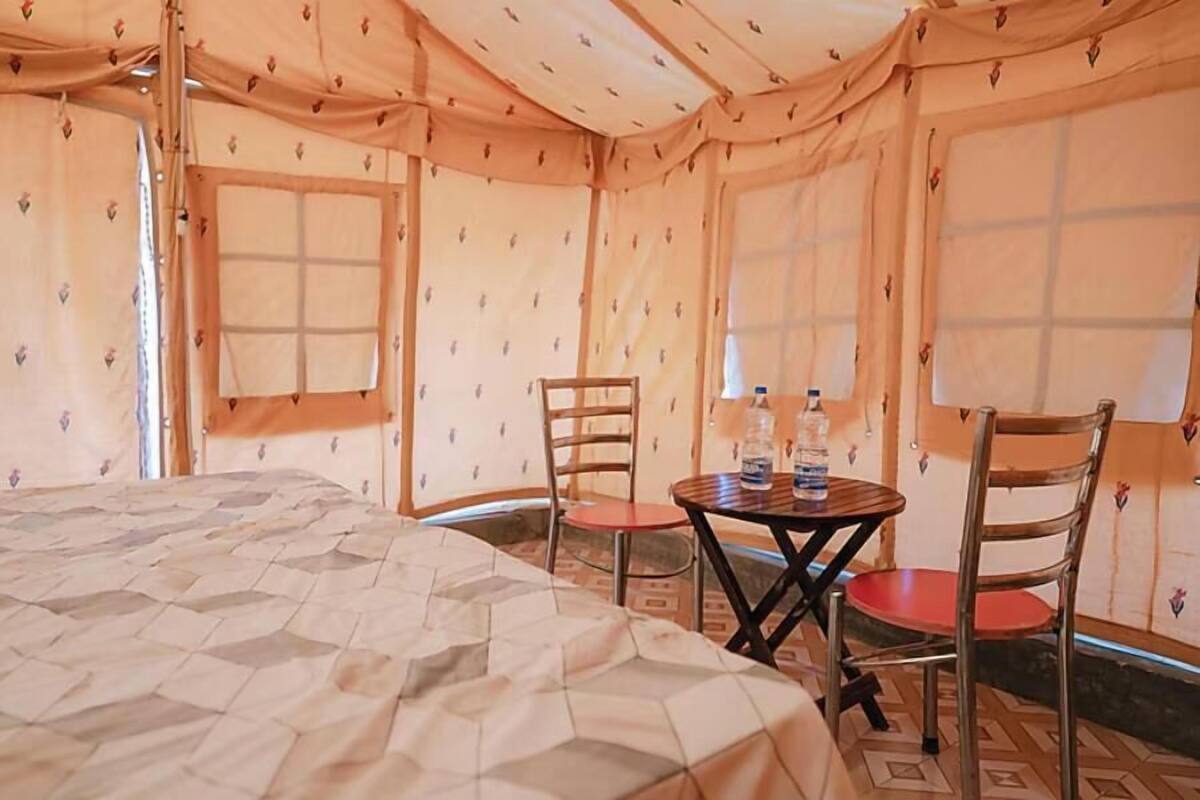 A tented room with a bed, two chairs, a small table with a bottle, and a window.