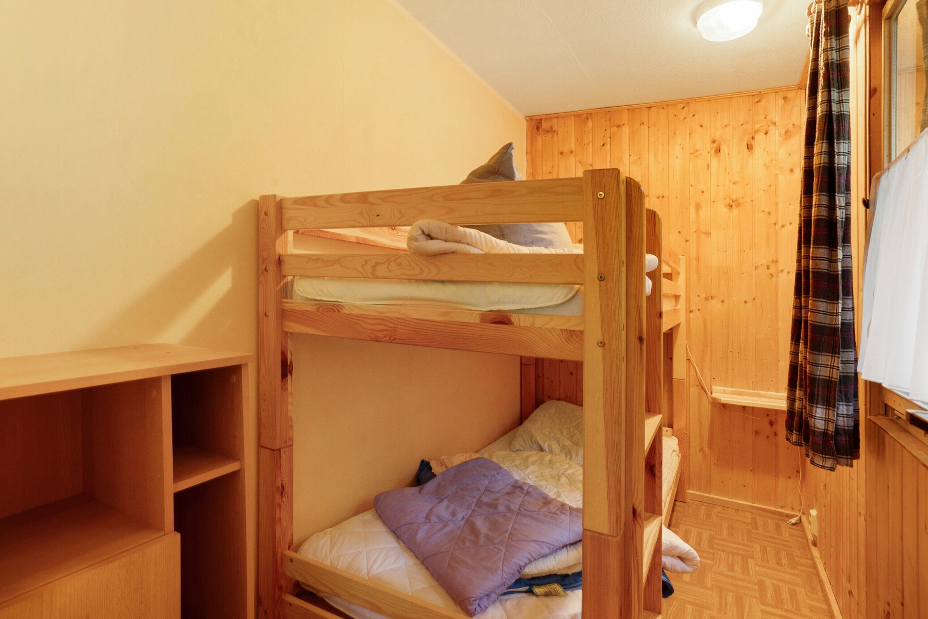 A bunk bed with a pillow and a blanket, wooden walls, a shelf, and a window with a curtain.