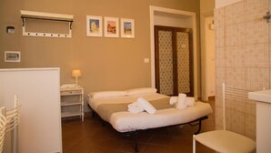Habitación de hotel con dos camas, una mesita con lámpara, zona de ducha y estante en la pared.