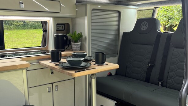 A compact RV interior with a kitchenette, seating area, and a window offering a view of greenery. - Glen Roamer VW Campervan – Explore Scotland in Comfort & Freedom (Dunblane)