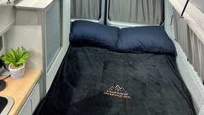 Een compact camperinterieur met een bed, een klein keukentje en een raam met gordijnen.
