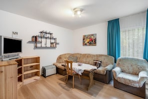 Un salon comprenant un canapé, deux fauteuils, une table basse, un téléviseur, une bibliothèque et un radiateur.