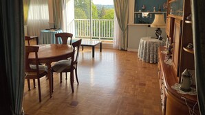 Une pièce spacieuse avec une table à manger en bois, des chaises, une petite table ronde avec une lampe, et une vue sur la verdure grâce à de grandes fenêtres.