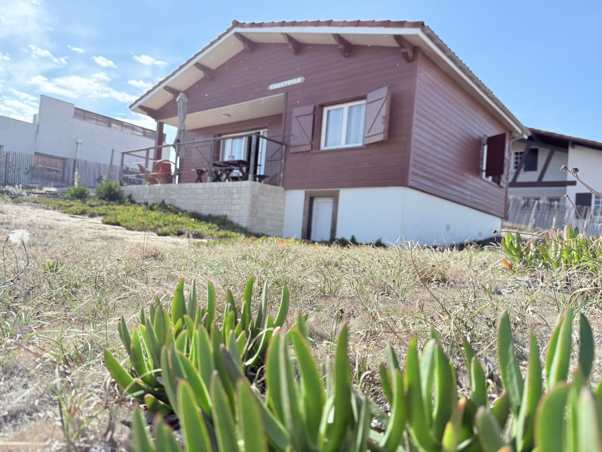 Una casa de dos pisos con balcón, rodeada de hierba seca y plantas.