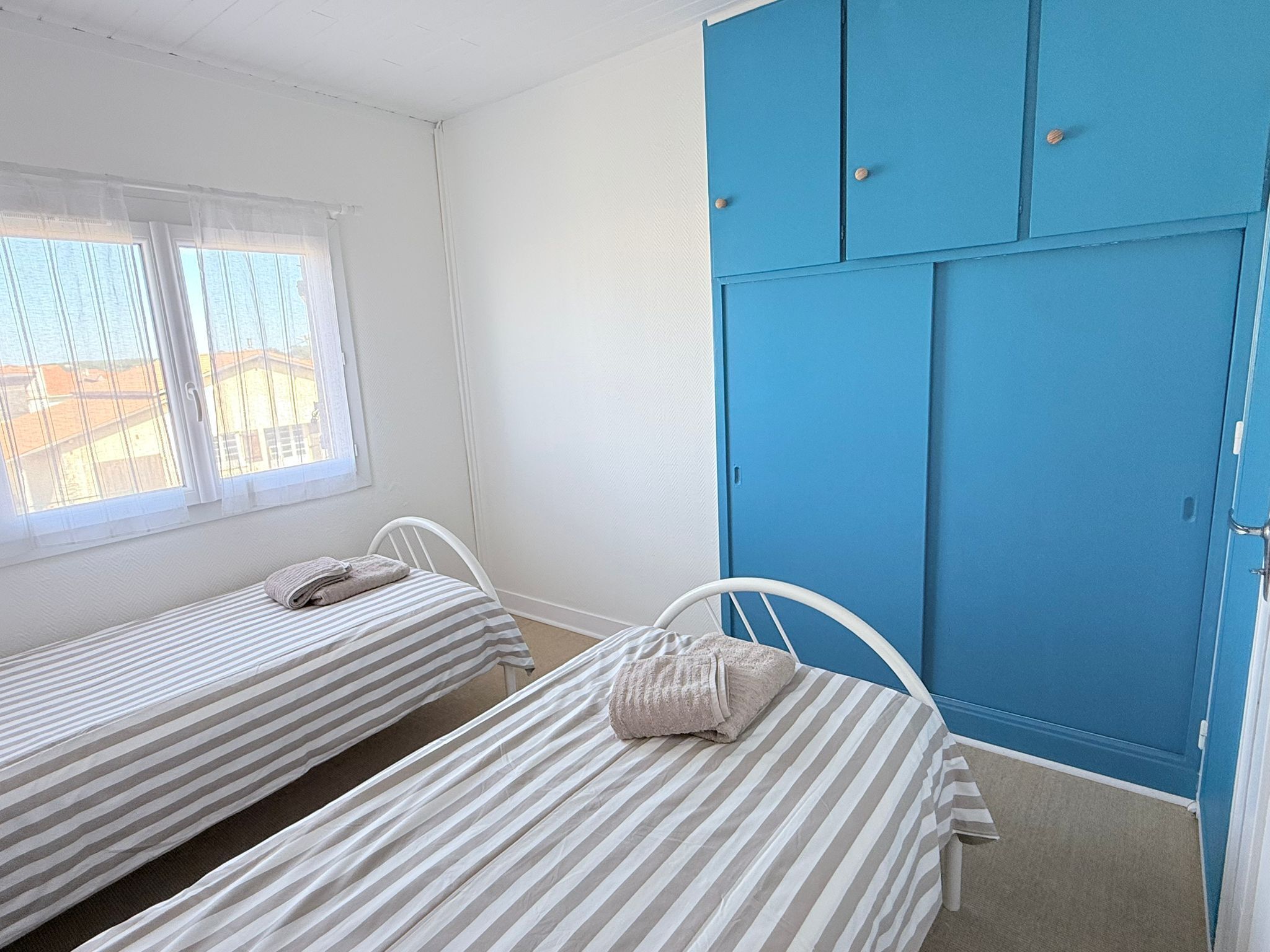 Une chambre avec deux lits simples, une fenêtre avec des rideaux légers et une armoire bleue.