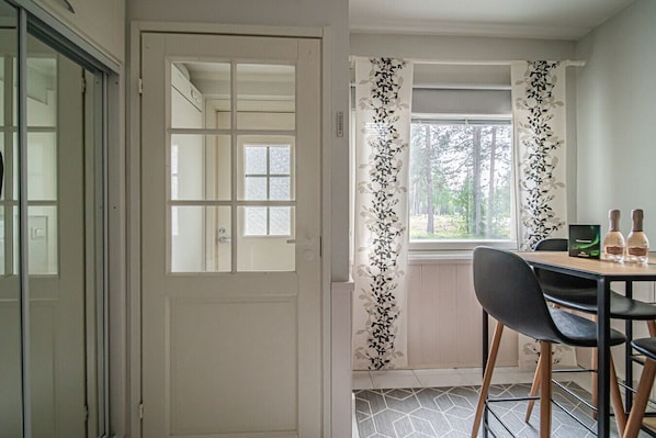 A room with a dining area, a white door with glass panels, and a window with a view of trees.