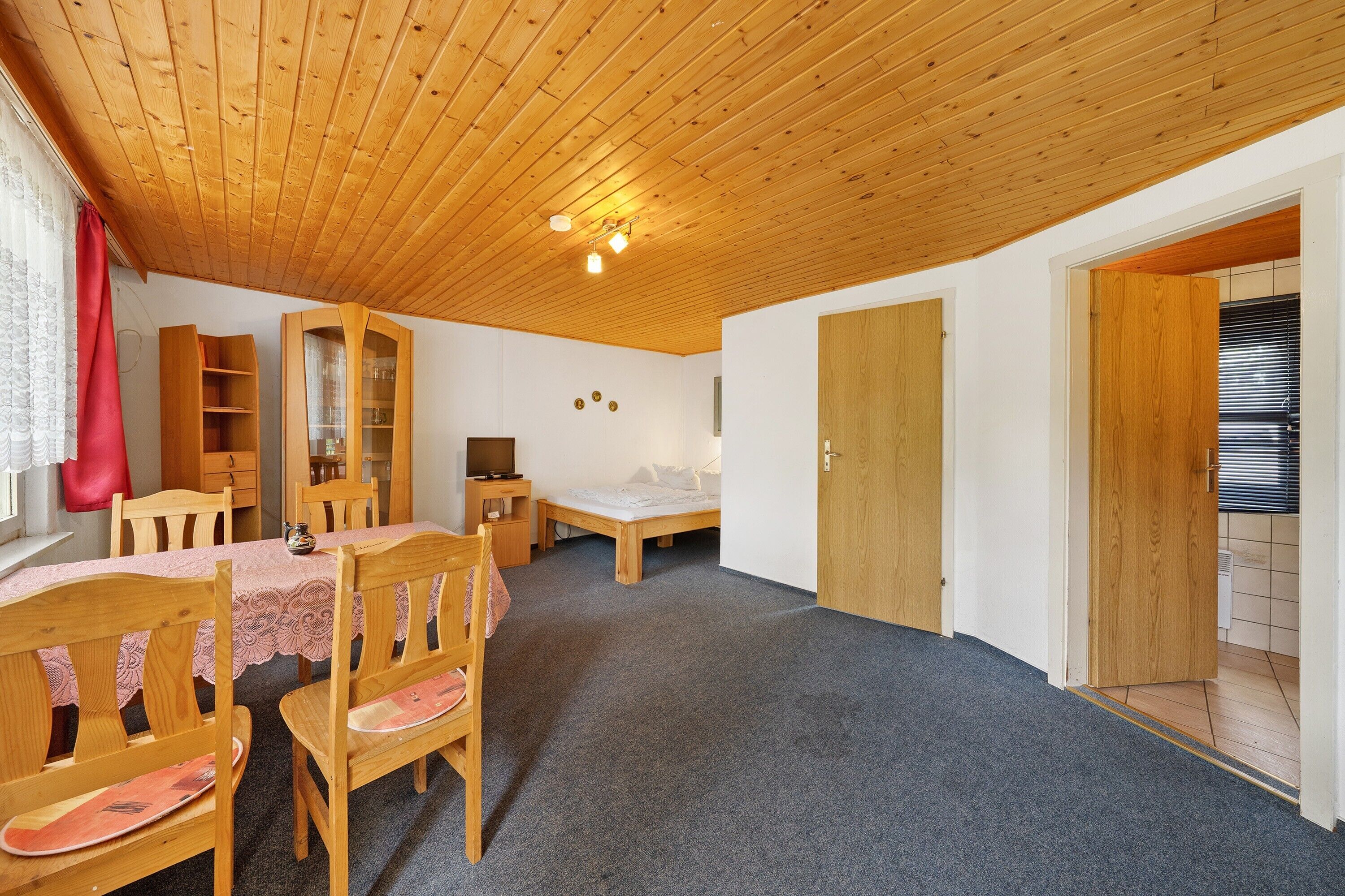 Ein Zimmer mit Holzboden, einem Esstisch mit Stühlen, einem Bett, einem Holzschrank und einer Tür zu einem weiteren Raum.