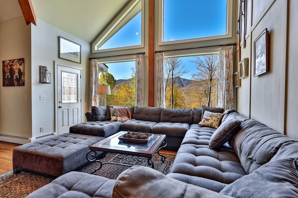 The vaulted living room includes sectional seating, tall windows, and mountain views.