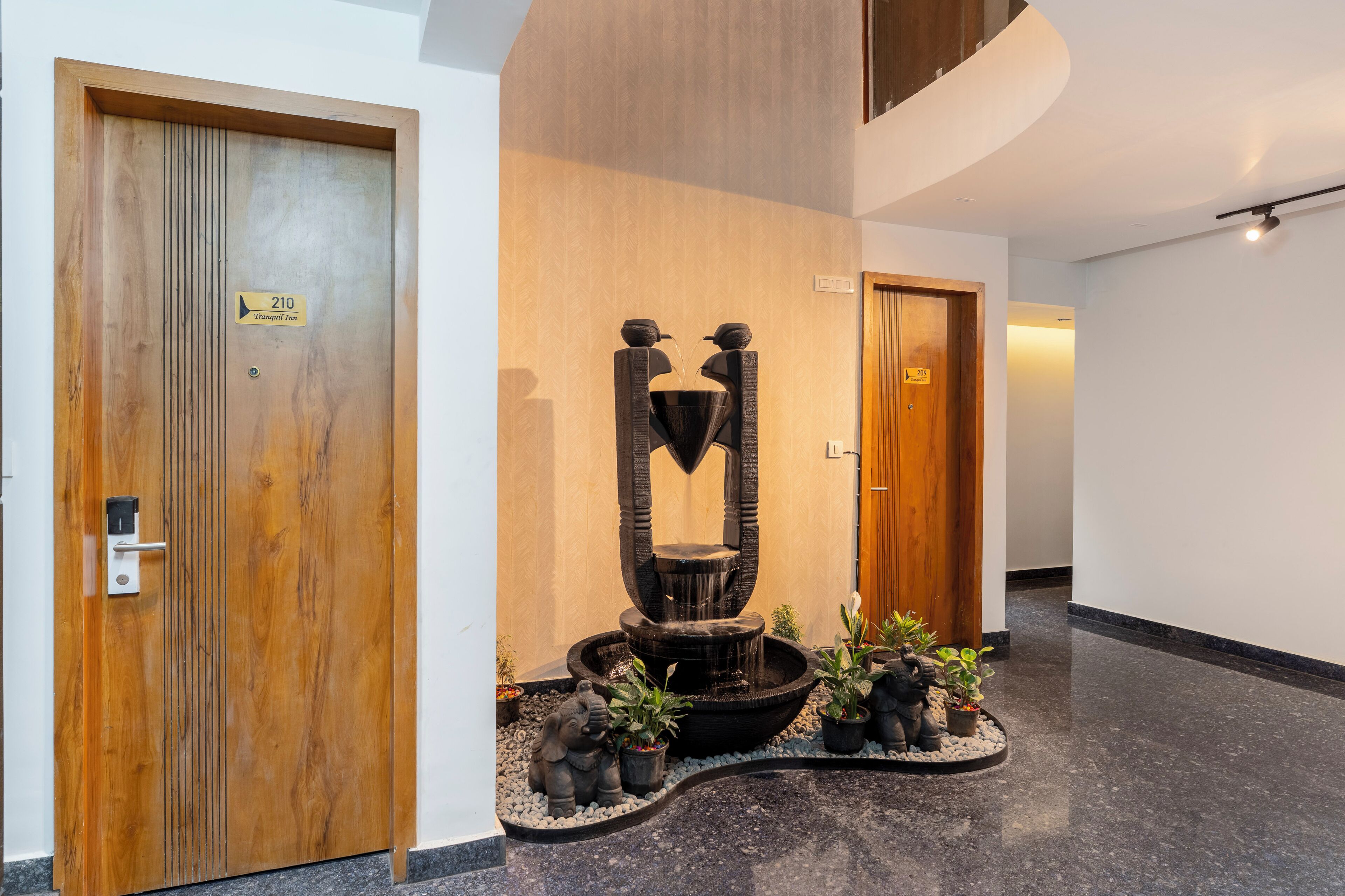 A modern hotel lobby with a wooden door labeled '210,' a water feature, and potted plants.