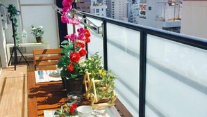 A balcony with a wooden table set for two, potted plants, and a view of the cityscape. - Hema Homestay Nihonbashi (Osaka)