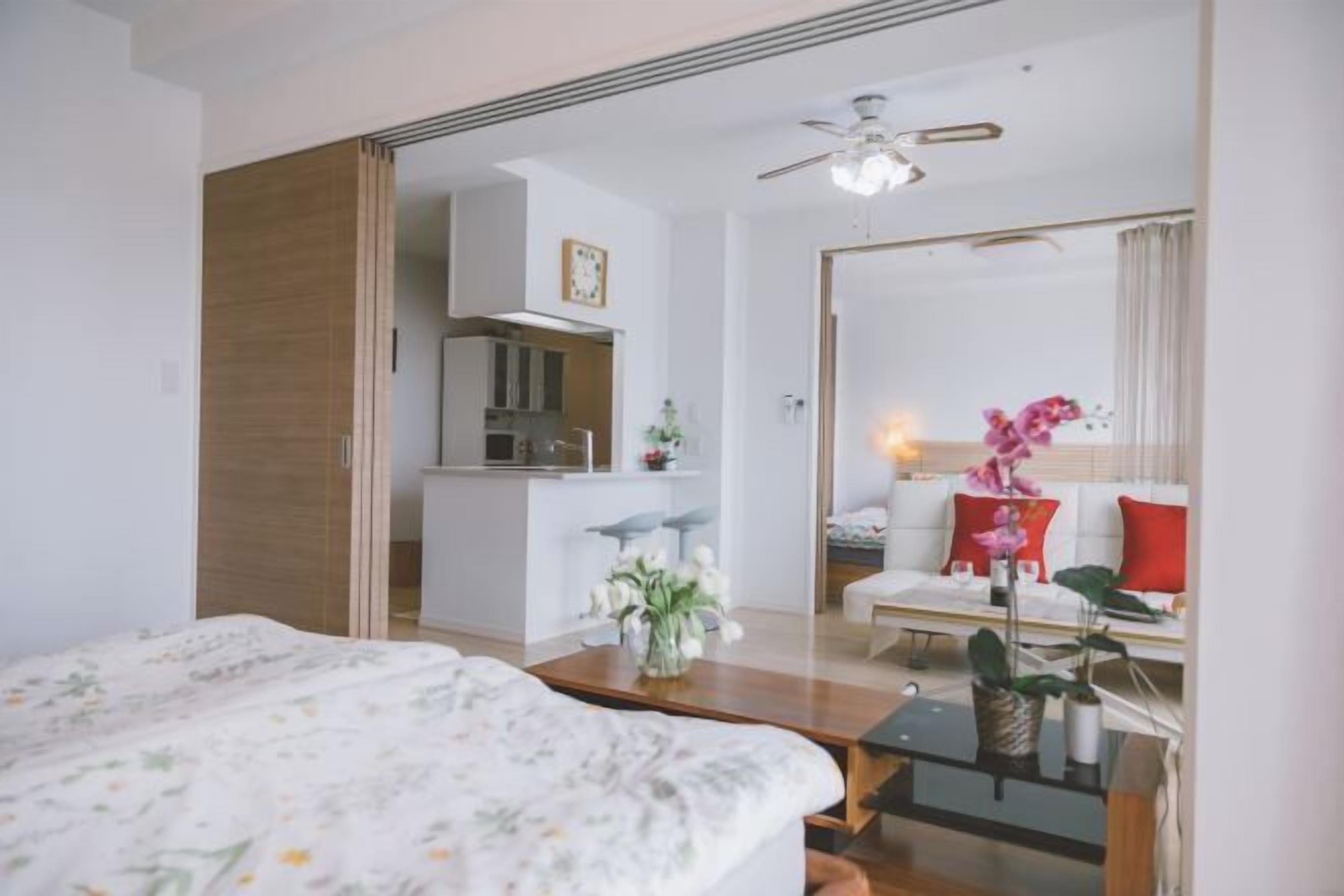 A modern hotel room with a bed, a small table with a vase of flowers, a sofa with cushions, and a kitchen area visible through an open door.
