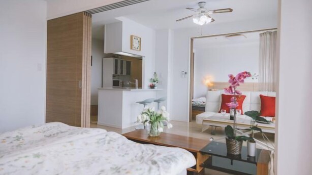 A modern hotel room with a bed, a small table with a vase of flowers, a sofa with cushions, and a kitchen area visible through an open door. - Hema Homestay Nihonbashi (Osaka)