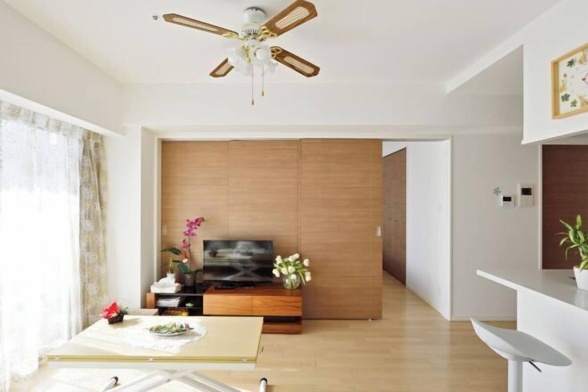 Una sala moderna con ventilador de techo, un televisor sobre un mueble de madera, una mesa de comedor con un jarrón de flores y una silla blanca.